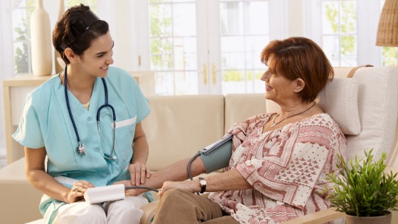 Medical Assistant sitting with an elderly female patient on couch in an assisted living facility, checking her blood pressure. MEDIjobs